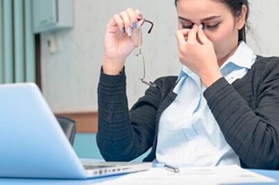 Imagem da notícia Imagem em formato horizontal. Uma mulher está sentada à mesa, diante de um notebook aberto. Ela usa camisa clara e blazer escuro. Com uma das mãos, segura um par de óculos; com a outra, toca a região entre os olhos. Sobre a mesa há papéis e uma caneta. O fundo mostra um ambiente interno com móveis claros.