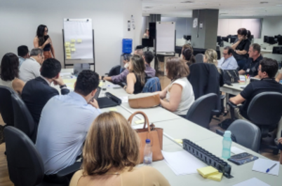 Imagem da notícia A imagem mostra um ambiente do Fórum Ruy Barbosa, onde um grupo de pessoas está reunido em uma atividade colaborativa. Há mesas dispostas no centro, ocupadas por participantes que assistem a uma apresentação em um flip chart, que tem post-its amarelos colados. No fundo, é possível observar um espaço amplo com mesas de trabalho e computadores. A iluminação é clara, típica de ambientes corporativos.