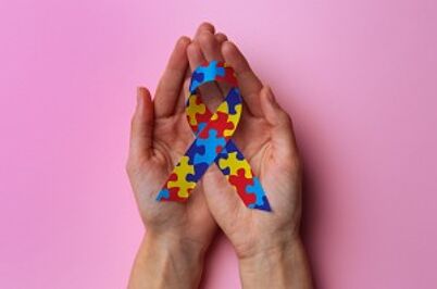 Imagem da notícia Duas mãos abertas segurando um laço com estampa de peças de quebra-cabeça coloridas (símbolo da conscientização do autismo) sobre um fundo rosa