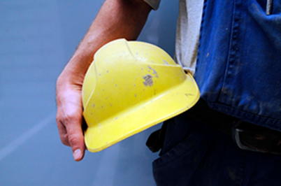 Imagem da notícia Imagem em formato horizontal. Um trabalhador segura um capacete de segurança amarelo com uma das mãos. O capacete apresenta marcas de uso e pequenas manchas. A pessoa veste roupa de trabalho azul, com bolso na altura do peito. Apenas o braço, parte do tronco e a mão são visíveis. O fundo é liso, em tom azul claro.