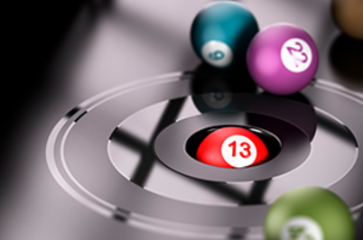 Imagem da notícia A imagem mostra bolas de bingo coloridas sobre uma superfície preta e brilhante, que lembra um alvo circular. No centro do alvo, há uma abertura circular onde uma bola vermelha com o número 13 está encaixada, destacando-se como o foco principal da imagem. Ao redor, outras bolas estão espalhadas: uma verde no canto inferior direito, uma azul e uma roxa ao fundo, ambas parcialmente desfocadas