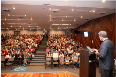 Imagem da notícia Imagem em formato horizontal. Um auditório amplo está cheio de pessoas sentadas em cadeiras organizadas em fileiras. No centro há uma escada que divide os assentos em dois blocos. À direita da imagem, em primeiro plano, um homem está em pé atrás de um púlpito, de costas para a câmera, segurando papéis. Ele usa terno escuro. À frente dele, o público está voltado para o palco. O ambiente possui paredes revestidas em madeira e teto com várias luzes embutidas.
