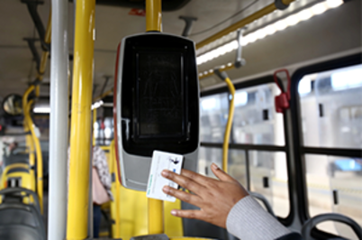 Imagem da notícia Mão de uma mulher segurando um cartão de passagem próximo a uma máquina de validar bilhetes no ônibus