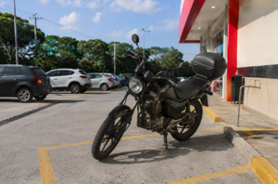 Imagem da notícia Moto estacionada em vaga ao lado de uma farmácia, em estacionamento pavimentado, com carros ao fundo e parte da fachada visível.