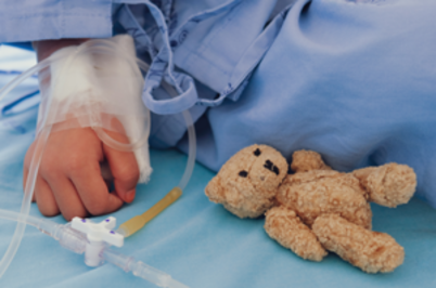 Imagem da notícia Criança com avental hospitalar, enquadramento em uma das mãos, que está com tubo hospitalar de silicone e tem um urso de pelúcia posicionado ao lado.