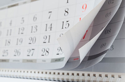 Imagem da notícia A imagem mostra um calendário de parede, com várias páginas sendo folheadas. As folhas estão parcialmente levantadas, revelando os dias e números dos meses seguintes. Os números estão impressos em preto, e alguns dias aparecem destacados em vermelho.