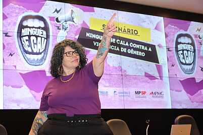 Imagem da notícia Uma mulher de óculos com cabelo cacheado, vestindo camiseta roxa, braço esquerdo tatuado erguido com o polegar para cima em frente a um telão com a frase "NINGUÉM SE CALA CONTRA A VIOLÊNCIA DE GÊNERO".