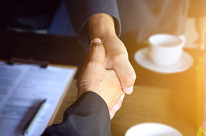 Imagem da notícia Imagem em formato horizontal. Duas pessoas estão apertando as mãos. Apenas os braços e as mãos são visíveis. Ambas usam roupas formais em tons escuros. O aperto de mãos ocorre sobre uma mesa. Sobre a mesa há papéis com texto impresso, uma pasta ou prancheta e, ao fundo, uma xícara branca sobre um pires. A iluminação é quente, com uma faixa de luz amarelada no lado direito da imagem.