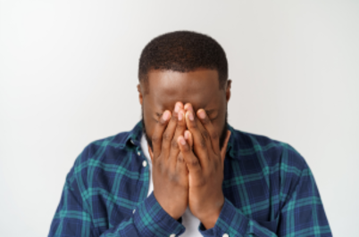 Imagem da notícia A imagem mostra um homem de pele escura com o rosto parcialmente coberto pelas mãos, como se estivesse triste, aflito ou cansado. 