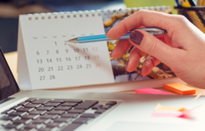 Imagem da notícia imagem foca em uma mão apontando datas em u m calendario de mesa com uma caneta. As unhas da mão estão pitnadas em uma cor vinho. Ao lado, visualiza-se o teclado de um notebook.