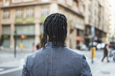 Imagem da notícia Homem negro, usando tranças no cabelo e de costas em uma rua, que está desfocada na imagem.