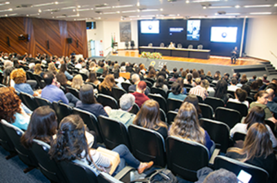 Imagem da notícia Imagem de um auditório amplo, visto a partir do fundo da sala. Há centenas de pessoas sentadas em fileiras de cadeiras estofadas escuras, voltadas para a frente. O público é diverso, com homens e mulheres adultos, usando roupas sociais e casuais, como camisas, blusas e blazers. À frente, há uma mesa longa elevada, onde várias pessoas estão sentadas, compondo uma mesa de autoridades e palestrantes. Atrás delas, um grande painel com telas e logotipos é projetado na parede, indicando a realização de um evento institucional.