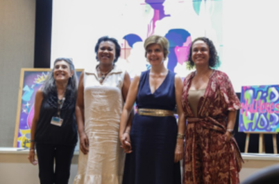Imagem da notícia Imagem em formato horizontal. Quatro mulheres estão lado a lado, de pé, posando para a foto. Elas estão de frente para a câmera e seguram as mãos entre si. Cada uma veste roupas diferentes: a primeira, à esquerda, usa roupa escura; a segunda usa vestido claro; a terceira usa vestido escuro sem mangas; e a quarta, à direita, usa vestido com estampa. Ao fundo, há um painel colorido com ilustrações e, à esquerda, um quadro apoiado. O ambiente parece ser interno, com iluminação artificial.
