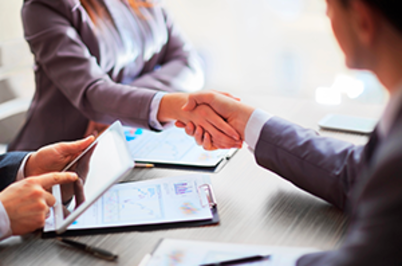Imagem da notícia Imagem em ambiente bem iluminado corporativo. Em primeiro plano, duas pessoas vestidas com roupas formais apertam as mãos sobre uma mesa, indicando um cumprimento profissional ou fechamento de acordo. Apenas os braços e as mãos são visíveis. À esquerda, outra pessoa, também com roupa social, segura uma prancheta com gráficos impressos e aponta para o documento com uma caneta. Sobre a mesa há papéis e um tablet. Ao fundo, levemente desfocadas, aparecem outras pessoas sentadas, sugerindo uma reunião.