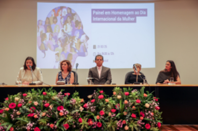 Imagem da notícia Cinco pessoas estão sentadas à mesa de um evento no auditório do Fórum Ruy Barbosa, com um arranjo de flores coloridas à frente. No centro da mesa, o presidente do Tribunal Regional do Trabalho da 2ª Região (TRT-2), desembargador Valdir Florindo, veste terno cinza e gravata, em posição de destaque. Ao fundo, é projetado um slide com a arte do evento e os dizeres “Painel em Homenagem ao Dia Internacional da Mulher”. À esquerda e à direita do desembargador, quatro mulheres participam da mesa, compondo o painel. O ambiente é iluminado e formal, com foco na celebração do Dia Internacional da Mulher.