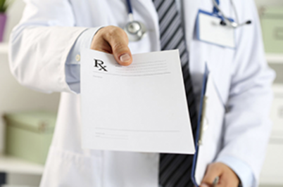 Imagem da notícia Imagem em formato horizontal. Em primeiro plano, uma pessoa veste jaleco branco e segura um papel com a mão estendida em direção à câmera. No papel há um símbolo no canto superior esquerdo semelhante a “Rx”. A pessoa usa camisa clara e gravata escura sob o jaleco. Um estetoscópio está pendurado no pescoço. Ao fundo, há um ambiente interno claro, com móveis desfocados.