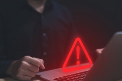 Imagem da notícia Em primeiro plano, há um notebook aberto sobre uma mesa, visto de lado. Na tela do computador aparece um símbolo luminoso em vermelho: um triângulo com um ponto de exclamação no centro, indicando alerta ou aviso. À esquerda da imagem, parcialmente visível, está uma pessoa vestindo uma camisa escura de mangas compridas. Apenas o tronco e uma das mãos podem ser vistos; a mão está próxima ao teclado, como se estivesse prestes a digitar ou interagir com o notebook. O ambiente é escuro, com iluminação baixa, o que destaca o brilho vermelho do símbolo de alerta na tela e cria uma atmosfera de atenção ou risco.
