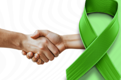 Imagem da notícia Imagem em formato horizontal. À esquerda, duas pessoas apertam as mãos; apenas as mãos e parte dos antebraços estão visíveis. À direita da imagem, aparece uma fita verde larga, dobrada em formato de laço. O fundo é claro, em tom branco ou cinza muito claro, com linhas suaves. A fita ocupa grande parte do lado direito da imagem.