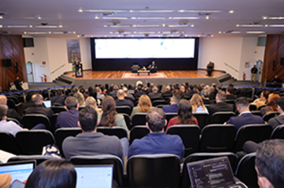 Imagem da notícia Plateia vista de costas durante fórum de governança; ao fundo, o palco principal