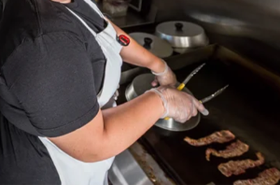 Imagem da notícia Pessoa vista parcialmente e de costas, usando avental branco sobre roupa escura e luvas descartáveis. Ela segura uma pinça e manipula alimentos sobre uma grelha ou chapa. À direita, há pedaços de carne sendo grelhados. Ao fundo, aparecem utensílios e panelas sobre uma superfície de cozinha.