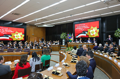 Imagem da notícia A imagem mostra o Tribunal Pleno do TRT-2 ocupado por homenageados(a) na Comenda e ao fundo, telão com arte específica do evento