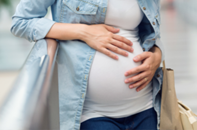 Imagem da notícia Imagem de uma pessoa grávida, vista do tronco até o quadril. Ela veste uma camiseta branca justa que evidencia a barriga de gravidez e uma camisa jeans aberta por cima. As duas mãos estão apoiadas sobre a barriga, em gesto de cuidado. A pessoa está encostada em um corrimão metálico. No braço direito, segura uma sacola.