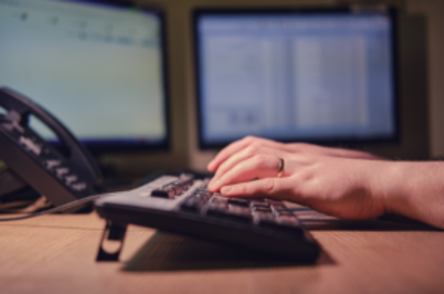 Imagem da notícia duas mãos digitam em um teclado de computador sobre uma mesa de madeira. Ao fundo, é possível visualizar um aparelho telefônico e dois monitores. O conteúdo dos monitores não está claro, mas parecem ser ferramentas de escritório.