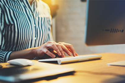 Imagem da notícia Imagem de meio corpo de mulher com camisa listada de azul-marinho teclando na frente do micro
