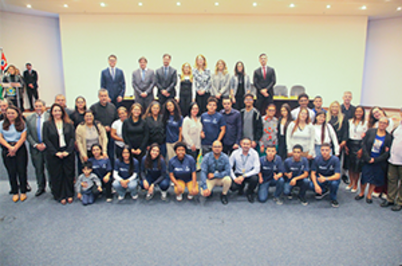 Imagem da notícia Cerca de 40 pessoas posam para foto oficial em auditório iluminado, durante a cerimônia de lançamento do Programa Adolescente-Jovem Aprendiz, no Fórum Ruy Barbosa. Na frente, jovens vestindo camisetas azuis e calças claras; atrás, adultos com trajes formais. O ambiente é solene e de celebração.