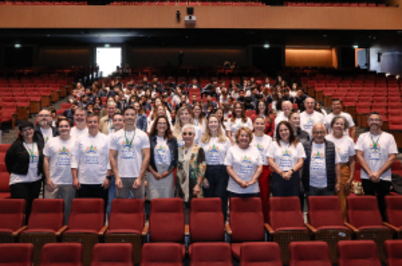 Imagem da notícia Um grande grupo posa para foto dentro de um auditório. Na frente, há diversas pessoas adultas alinhadas, muitas usando camisetas brancas com estampa de um evento. Ao fundo, ocupando as cadeiras vermelhas do teatro, estão dezenas de estudantes e participantes. O ambiente é amplo, com palco atrás do grupo e iluminação interna suave.