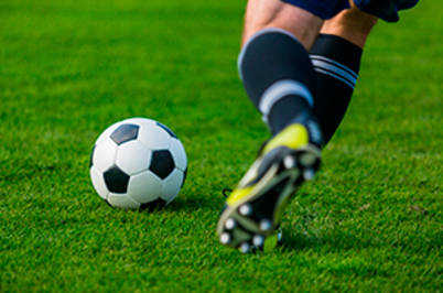 Imagem da notícia Parte inferior das pernas de um jogador de futebol correndo atrás de uma bola em campo gramado