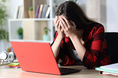 Imagem da notícia A imagem mostra uma mulher sentada à frente de um notebook vermelho em uma mesa. Ela está com as duas mãos cobrindo o rosto, aparentando estar angustiada ou frustrada. Veste uma camisa xadrez vermelha e preta e tem cabelos castanhos, lisos e compridos. Ao fundo, vê-se uma estante com livros e objetos decorativos, indicando um ambiente de escritório. Sobre a mesa, além do notebook, há fones de ouvido brancos e alguns cadernos ou blocos de anotações.