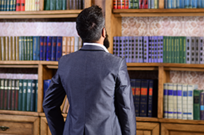 Imagem da notícia A imagem mostra um homem de costas, vestindo um terno escuro e uma camisa branca. Ele tem cabelos curtos e barba cheia. O homem está em frente a uma grande estante de madeira repleta de livros organizados em diferentes cores e tamanhos. A estante tem um estilo clássico, e o ambiente sugere um escritório ou biblioteca