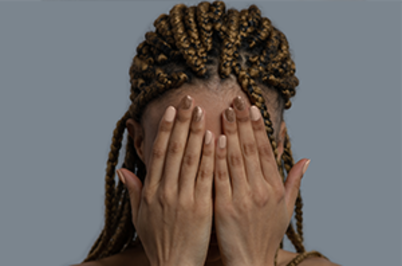 Imagem da notícia A imagem mostra uma pessoa de pele negra com tranças presas à cabeça, ocupando o centro do enquadramento. Ela está de frente, mas com o rosto completamente coberto pelas duas mãos, como se estivesse constrangida. Os dedos estão estendidos, tocando-se levemente, escondendo todos os traços faciais. O fundo é liso e acinzentado, sem objetos visíveis, dando destaque à figura em primeiro plano.