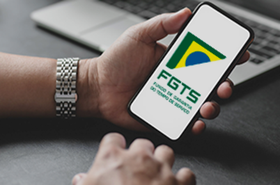 Imagem da notícia Imagem em formato horizontal. Uma pessoa segura um smartphone com a tela voltada para cima. Na tela aparece o logotipo do FGTS, com as letras “FGTS” em verde e um elemento gráfico nas cores verde, amarelo e azul, remetendo à bandeira do Brasil. A mão que segura o aparelho usa um relógio metálico prateado no pulso. Ao fundo, sobre uma mesa escura, há um notebook parcialmente visível.