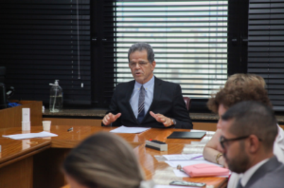 Imagem da notícia Em uma sala de reuniões iluminada por luz natural que entra através de persianas escuras, Dr. Valdir Florindo, de terno escuro e gravata azul fala enquanto gesticula com as mãos sobre a mesa. Ele está sentado à cabeceira, aparentando ser a pessoa que conduz a reunião. À frente dele, há papéis, um copo descartável e uma garrafa de vidro com água. Outras pessoas, parcialmente visíveis, estão sentadas ao redor da mesa, prestando atenção.