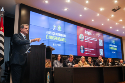 Imagem da notícia Imagem em formato horizontal. Em um auditório, o desembargador-presidente Valdir Florindo está em pé atrás de um púlpito, falando ao microfone. Ele usa terno escuro, camisa clara e gravata. À direita do púlpito há uma mesa longa onde várias pessoas estão sentadas lado a lado. Ao fundo, um grande painel digital ocupa a parede, dividido em duas partes com fundo azul e vermelho e textos projetados. No canto esquerdo da imagem há bandeiras em mastros. O teto do auditório tem várias luzes embutidas.