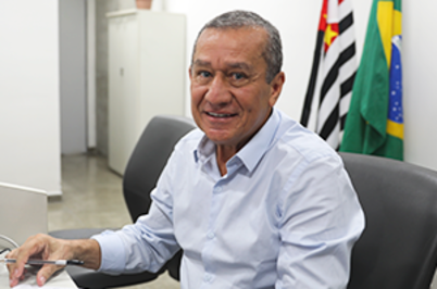 Imagem da notícia Uma fotografia em plano médio e enquadramento horizontal que mostra um homem de meia-idade sentado em um escritório. Ele está posicionado no centro da imagem, voltado para a câmera com um sorriso suave. Ao fundo, à direita, estão as bandeiras do estado de São Paulo e do Brasil.