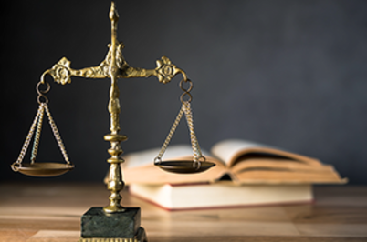 Imagem da notícia Imagem mostra uma balança de contrapeso que representa a justiça e um libro aberto, representando a lei. Os objetos estão sobre uma mesa de madeira.