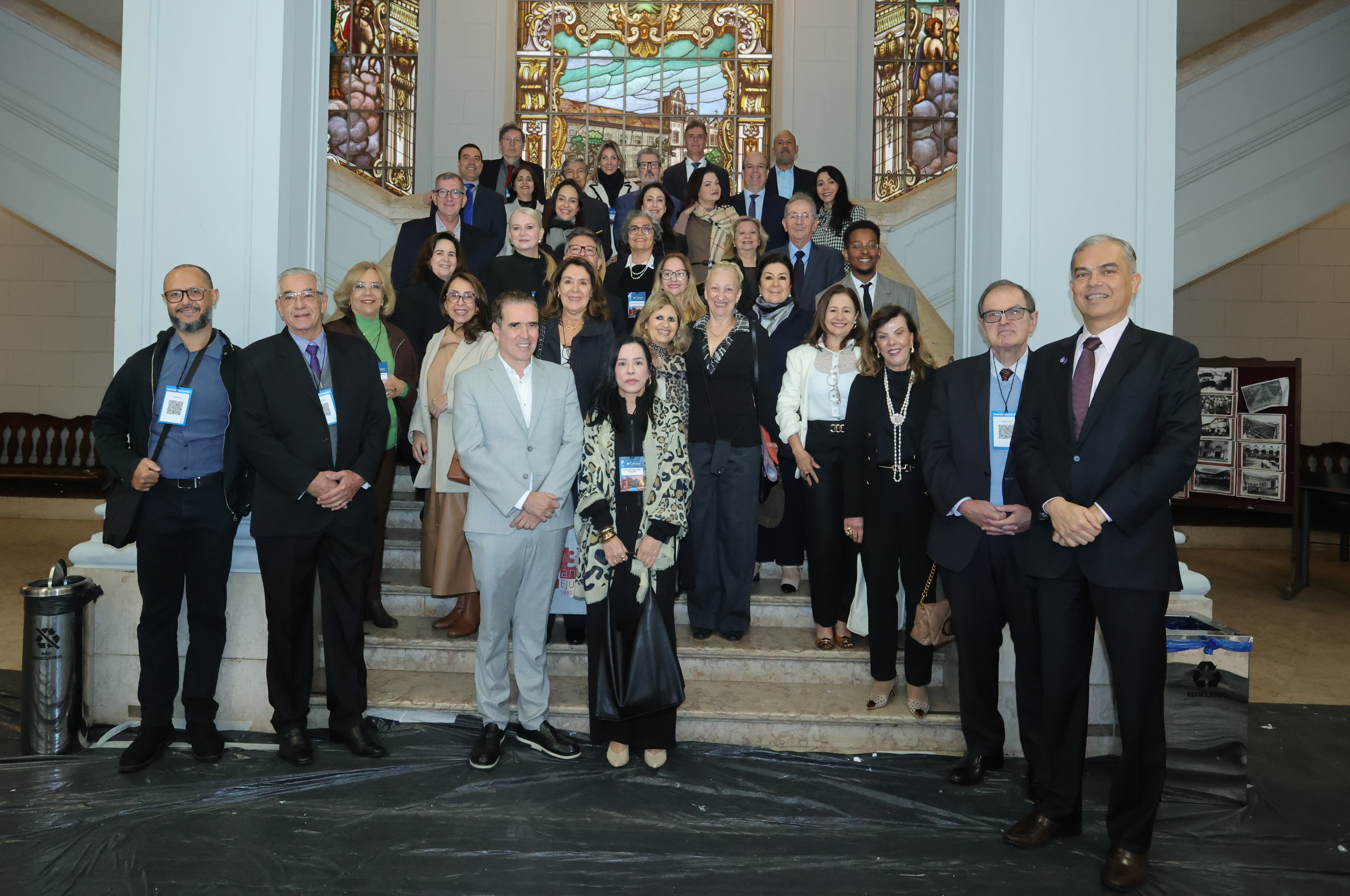 Imagem da Notícia Imagem em formato horizontal. Um grupo de pessoas está reunido em uma escadaria, posando para uma fotografia. Há pessoas em várias fileiras, algumas em pé nos degraus superiores e outras na parte inferior. Na primeira fileira, aparecem homens e mulheres usando roupas sociais, com ternos, blazers e vestidos. Ao fundo, há um grande vitral colorido emoldurado por uma estrutura clara. Nas laterais da escadaria, há corrimãos escuros e paredes claras.
