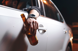 Imagem da Notícia imagem foca na janela de um carro e no braço de uma pessoa para fora da janela do veículo, segurando uma garrafa long neck de cerveja