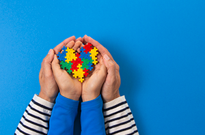 Imagem da Notícia Mãos de criança segurando um coração montado com peças de quebra-cabeças, envoltas por mão de adulto, em fundo azul