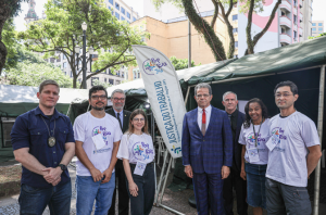 Imagem da Notícia Imagem em ambiente externo, durante o dia. Sete pessoas estão alinhadas, posando para foto. Ao centro, um homem veste terno azul e gravata vermelha. Os demais usam roupas casuais, com predominância de camisetas claras estampadas.  Ao fundo, há árvores, uma estrutura tipo tenda à direita e parte de um edifício ao fundo. Iluminação natural, com sombras suaves. Todos estão voltados para a câmera.