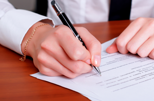 Imagem da Notícia Imagem em close. Sobre uma mesa de madeira clara, vê-se um documento impresso em papel branco. Duas mãos humanas aparecem em primeiro plano. A mão direita segura uma caneta preta e está escrevendo ou assinando o documento. A mão esquerda apoia o papel, mantendo-o firme. A pessoa veste uma camisa branca de manga comprida; no pulso esquerdo há uma pulseira fina dourada. O texto do documento não é legível.