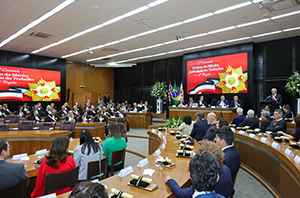 Imagem da Notícia A imagem mostra o Tribunal Pleno do TRT-2 ocupado por homenageados(a) na Comenda e ao fundo, telão com arte específica do evento