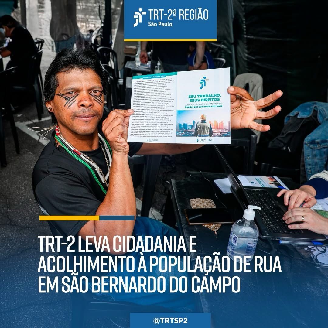  A imagem mostra um homem com traços indígenas, cabelos pretos lisos e curtos, usando colares, e sentado em um espaço coberto. Ele está sorrindo levemente e segura com as duas mãos um folheto aberto do TRT-2 com a frase: "Seu trabalho, seus direitos – Direitos que caminham com você".