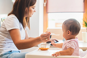Imagem da Notícia Imagem de moça de camiseta branca dando comida para bebê no cadeirão, ambos vistos de perfil