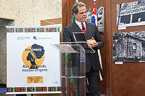 Imagem da Notícia Dr. Valdir Florindo discursa em púlpito para abrir a exposição de fotografia. Atrás dele, as bandeiras do Brasil e do estado de São Paulo e algumas das fotos da exposição.