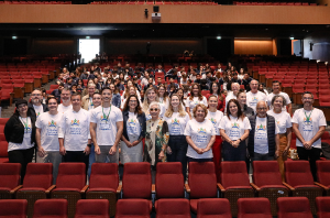 Imagem da Notícia Um grande grupo posa para foto dentro de um auditório. Na frente, há diversas pessoas adultas alinhadas, muitas usando camisetas brancas com estampa de um evento. Ao fundo, ocupando as cadeiras vermelhas do teatro, estão dezenas de estudantes e participantes. O ambiente é amplo, com palco atrás do grupo e iluminação interna suave.