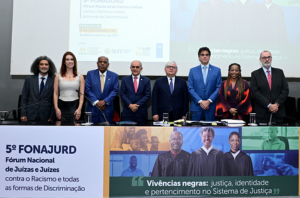 Ir para Imagem da Notícia Mesa de abertura do encontro mencionado na notícia, com a presença de ministros do STJ e outros magistrados. Na bancada, um banner tem o texto "5º Fonajurd"